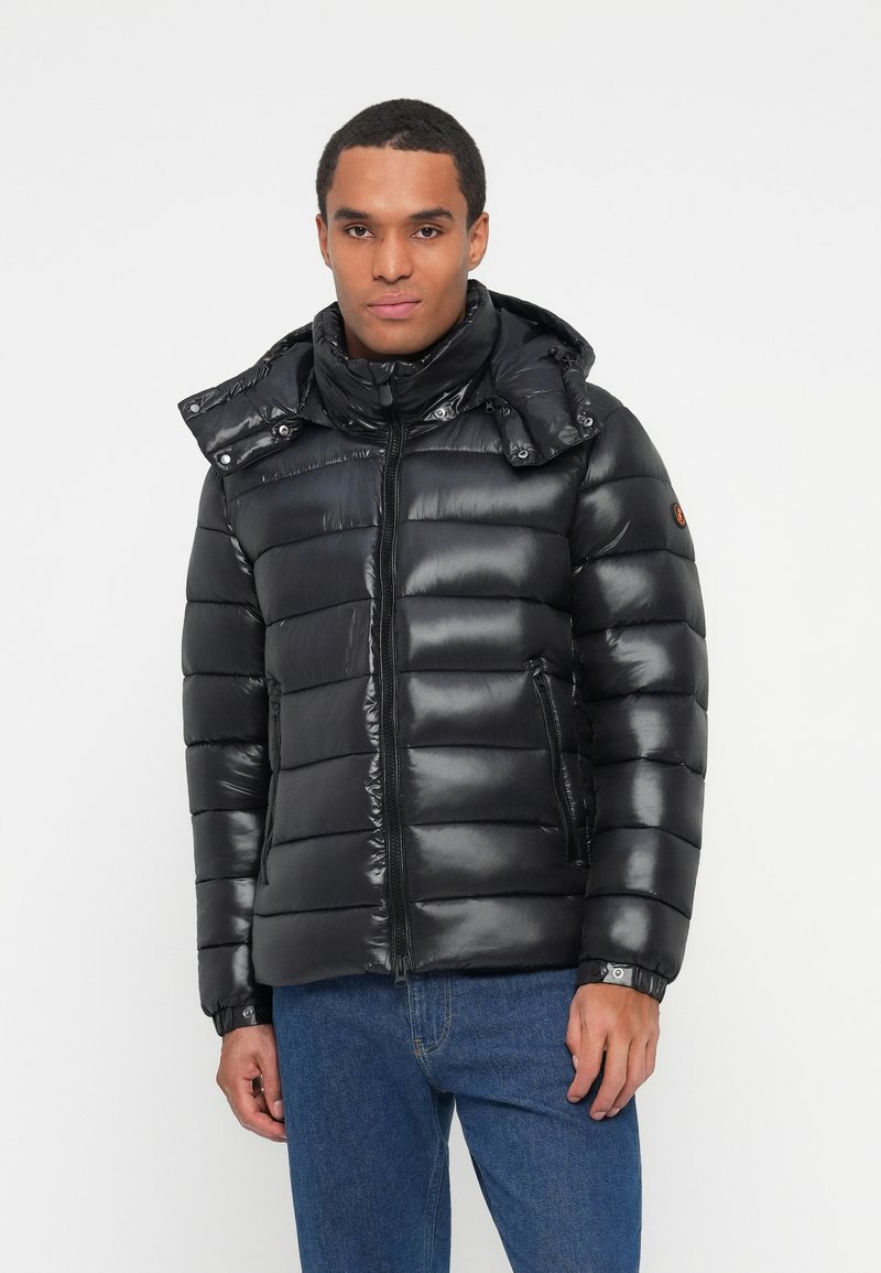 Uomo che indossa un giubbotto imbottito lucido nero con colletto alto, tasche con zip e jeans blu su uno sfondo bianco.