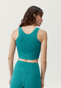 Tyrkysová sportovní podprsenka s upnutým střihem, ozdobená perforovanými detaily na zadní straně a zkrácenou délkou, vyrobena z elastického materiálu.