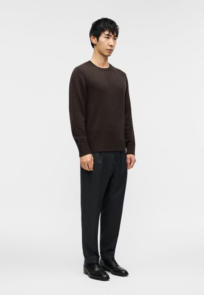 Καφέ πλεκτό πουλόβερ με στρογγυλή λαιμόκοψη, συνδυασμένο με σκούρο παντελόνι με ρίγες και μαύρα επίσημα παπούτσια· λεία υφή και ραμμένη εφαρμογή.