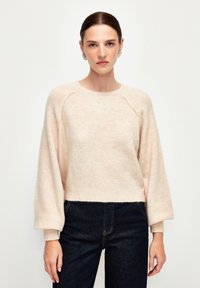 Hellrosa, kurzes Sweatshirt mit langen Ballonärmeln. Hergestellt aus weichem, flauschigem Material mit gerippten Bündchen und Ausschnitt. Kombiniert mit dunkelblauer Jeans.