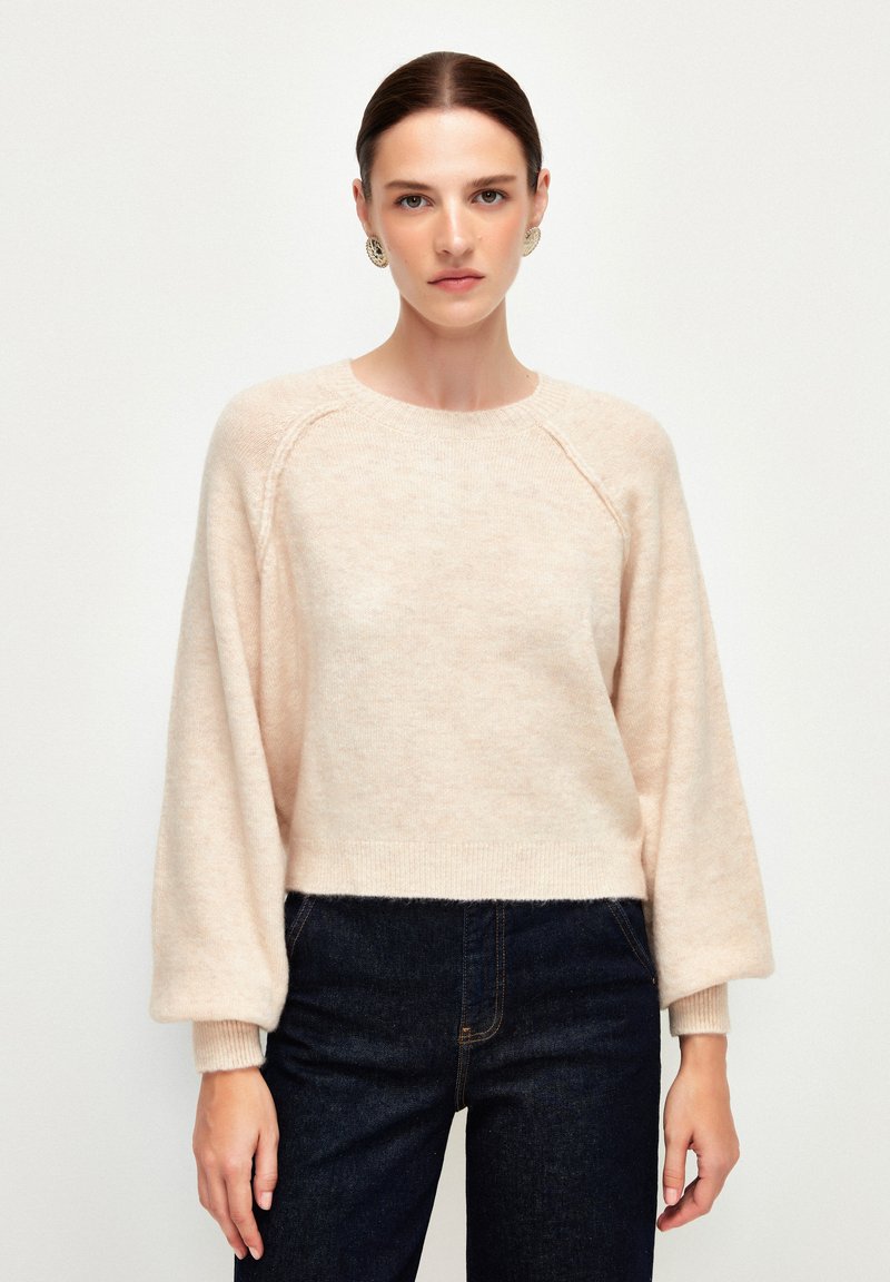 Hellrosa, kurzes Sweatshirt mit langen Ballonärmeln. Hergestellt aus weichem, flauschigem Material mit gerippten Bündchen und Ausschnitt. Kombiniert mit dunkelblauer Jeans.