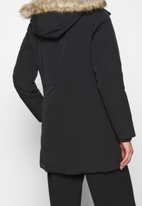 Back view of person wearing black winter coat with fur-trimmed hood and black pants, standing against plain light background.