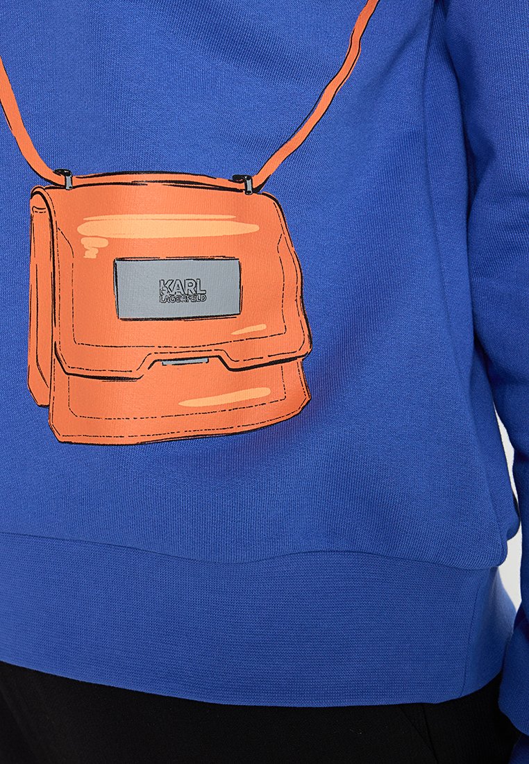 Suéter azul con una bolsa de hombro ilustrada en naranja en la parte frontal, con una placa de logo plateada. Tejido texturizado con puños acanalados.