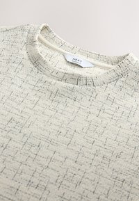 Close-up of a light beige textured sweater with a round neckline and black irregular grid pattern. Label reads "NEXT Relaxed Fit."