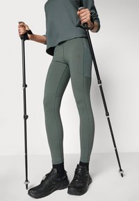 Olíva zöld sport leggings, rugalmas anyagból készült, varrás részletekkel. Fekete túracipővel és túrabotokkal párosítva.