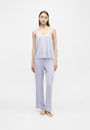 Jeune femme debout pieds nus portant un caraco sans manches lavande clair et un pantalon de pyjama ample assorti sur un fond blanc uni.