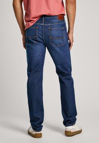 Jeans azul para hombre con un diseño de pierna recta, bolsillos traseros y un parche de cuero marrón en la cinturilla. El material parece ser denim.
