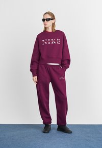 Bordo Nike sweatshirt z belim logotipom, v kombinaciji s ujemajočimi se trenirkami. Komplet ima sproščen kroj, mehak material in elastičen pas.