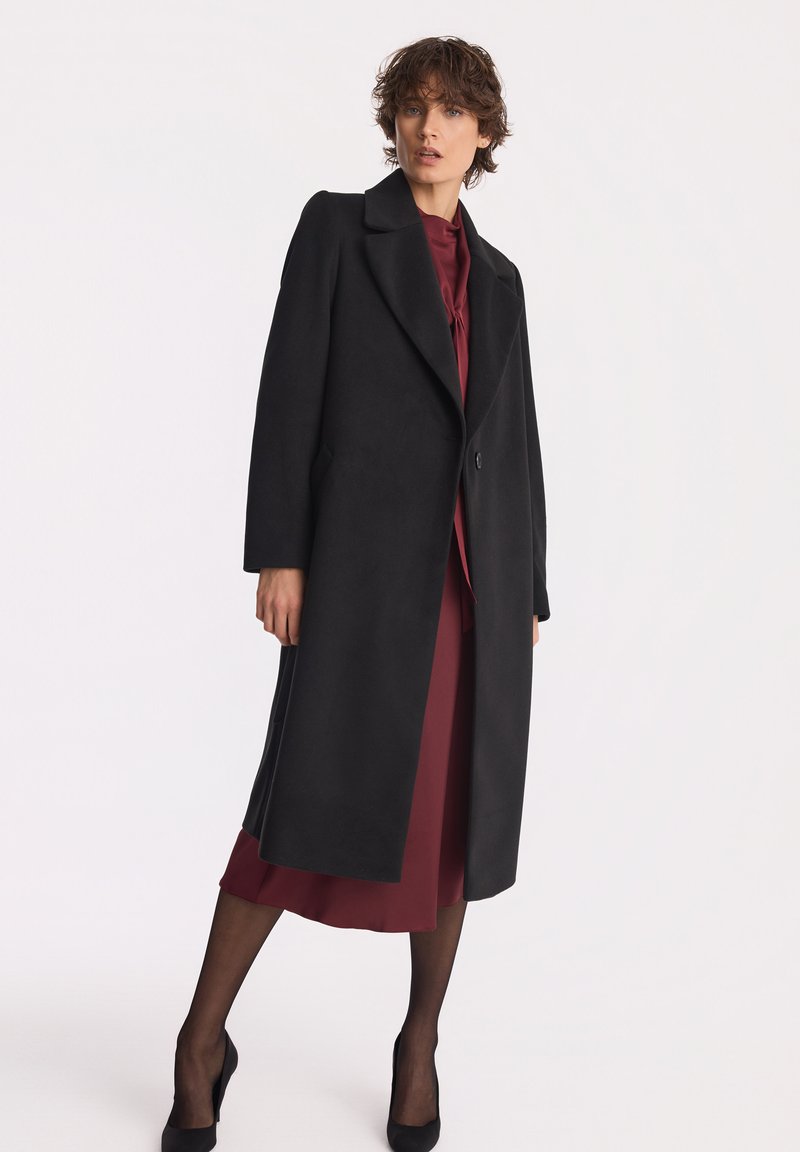 Kobieta w długim czarnym płaszczu, nosząca burgundową sukienkę, przejrzyste czarne rajstopy i czarne szpilki, stojąca na jednolitym jasnym tle.