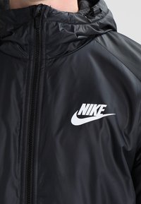 Svart Nike-jacka med dragkedja, huva och en vit Nike-logotyp på bröstet. Materialet ser slitstarkt och lätt ut.