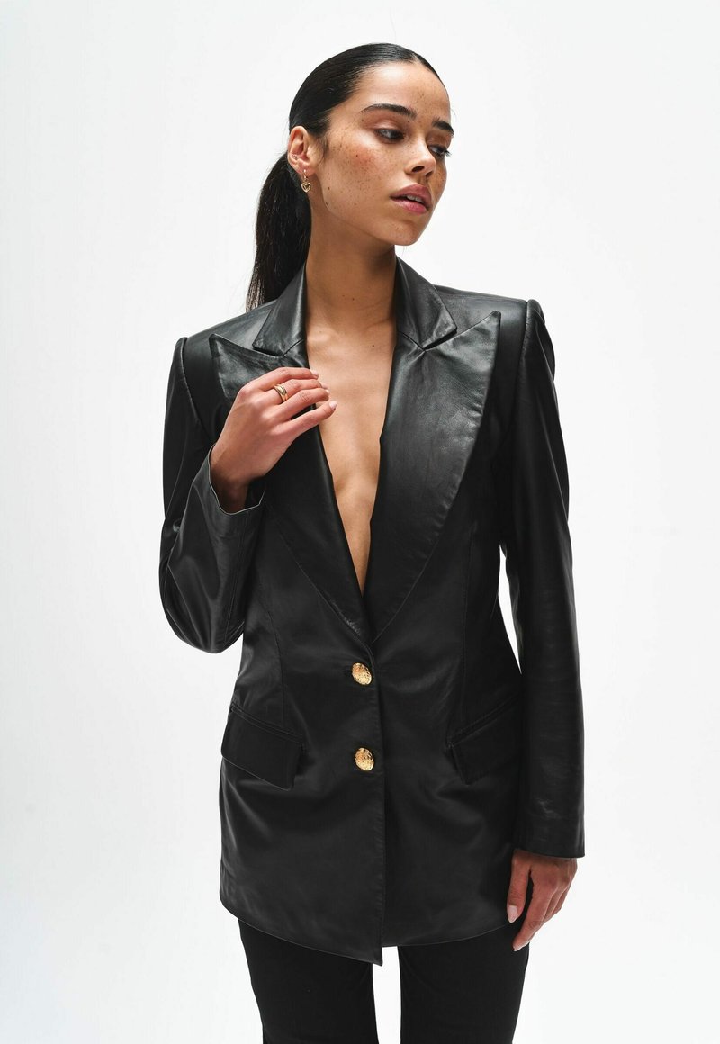 Kobieta o ciemnych włosach związanych w niski kucyk, ubrana w czarną skórzaną marynarkę z złotymi guzikami, pozująca na jednolitym białym tle.