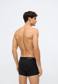 Mladý muž stojí s otočeným zády, oblečený v černých boxerách Calvin Klein na jednobarevném světlém pozadí.
