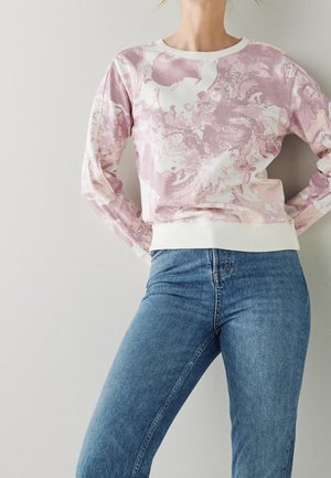 Osoba ubrana w różowo-biały sweter z marmurkowym wzorem i długimi rękawami, zestawiony z niebieskimi dżinsami z wysokim stanem, stojąca na prostym tle.