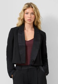 Blonda sieviete ar viļņainiem matiem, ģērbta melnā �īsā žaketē virs tumši sarkanas blūzes un atbilstošiem melniem augstiem biksēm, rokas kabatās.