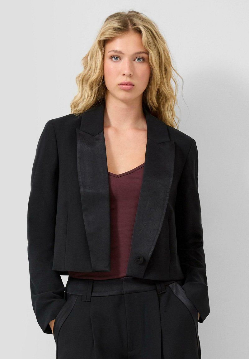 Blonda sieviete ar viļņainiem matiem, ģērbta melnā īsā žaketē virs tumši sarkanas blūzes un atbilstošiem melniem augstiem biksēm, rokas kabatās.