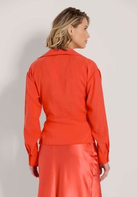 Rote Bluse mit langen Ärmeln und Kragen; hat eine taillierte Taille, glatte Textur und lose Ärmel mit geknöpften Manschetten.