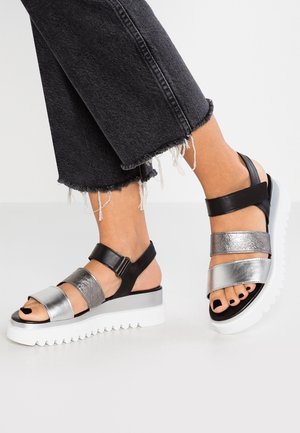 Fødder iført sølv- og sort tykke rem-sandaler med hvide platformsåler, kombineret med sorte jeans med opsmøgede kanter.