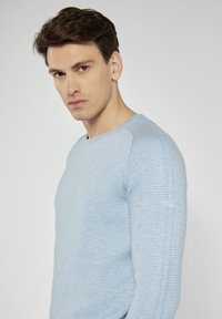 Helles blaues strukturiertes Sweatshirt mit geripptem Design, Rundhalsausschnitt und kleinem Etikett-Detail am linken Ärmel. Figurbetonte Silhouette.
