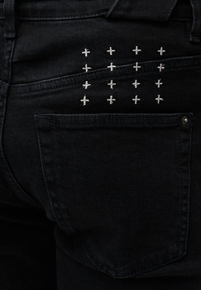 Mustad teksapüksid, mille tagataskul on valgete ristpistesdetailidega ruudustikmustris kaunistus.