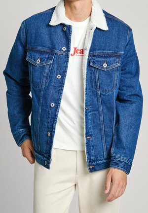 Veste en jean bleu moyen avec col en fausse fourrure, deux poches poitrine, boutons en métal argenté, et surpiqûres orange contrastantes, portée sur une chemise blanche.