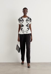 Witte top met zwarte bloemige borduursels, korte mouwen en een peplum zoom; gecombineerd met zwarte broek en een zwarte handtas. Platte schoenen.