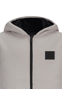 Felpa grigia chiara con chiusura a zip nera e morbida fodera nera. Presenta una patch logo quadrata sul lato sinistro del petto. Il tessuto è liscio ed elastico.
