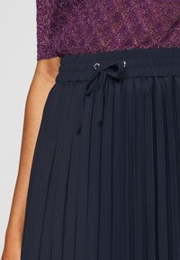 Gonna plissettata blu navy con vita elasticizzata, dotata di un cordino e una superficie liscia, abbinata a un top viola glitterato.