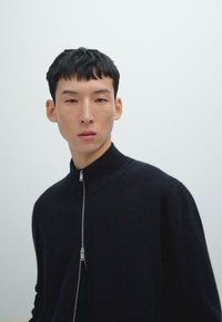 Junger Mann mit kurzen schwarzen Haaren, der einen schwarzen Reißverschluss-Pullover vor einem einfarbigen, hellgrauen Hintergrund trägt.