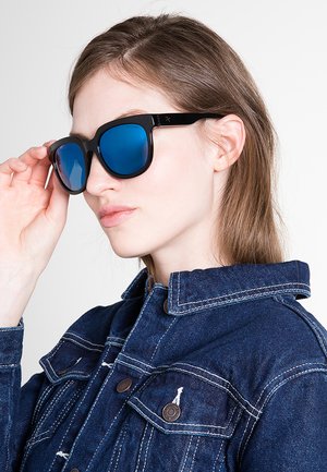 Giovane donna con giacca di jeans blu regola degli occhiali da sole con montatura nera e lenti blu riflettenti su uno sfondo semplice.