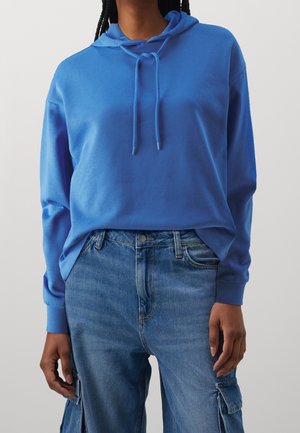 Person wearing a bright blue hoodie with drawstrings and high-waisted blue denim cargo jeans, standing against a plain background.