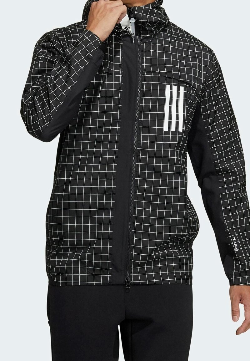 Musta takki, jossa on valkoinen ruutukuosi, edessä vetoketju, rinnan kohdalla kolme valkoista nauhaa, hupullinen muotoilu, sileä pinta ja joustavat hihansuut.