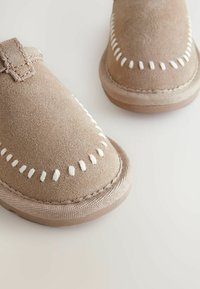 Jasnobeżowe zamszowe buty wsuwane z białym przeszyciem wzdłuż górnej krawędzi oraz teksturowaną, beżową gumową podeszwą. Płaski fason z okrągłym noskiem.