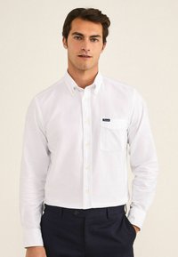 Camisa blanca de botones fabricada en algodón, con un bolsillo en el pecho y un pequeño parche con el logotipo. Mangas largas con puños abotonados y un cuello clásico.