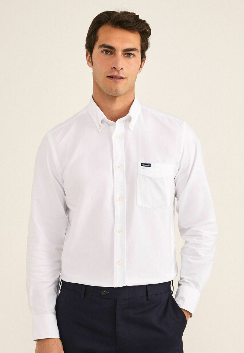 Camisa blanca de botones fabricada en algodón, con un bolsillo en el pecho y un pequeño parche con el logotipo. Mangas largas con puños abotonados y un cuello clásico.