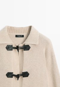 Beige knitted cardigan featuring a ribbed collar and two black toggle closures. The fabric appears soft with a textured finish.