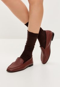 Barna bőr slip-on cipők sima felülettel, enyhén hegyes orral és alacsony fekete sarokkal, sötét bordázott zoknival párosítva.