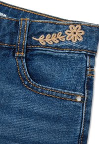 Dunkelblaue Denim-Jeans haben eine Fronttasche, goldene Nähte und beiges florales Stickmuster mit Blättern am Bund.