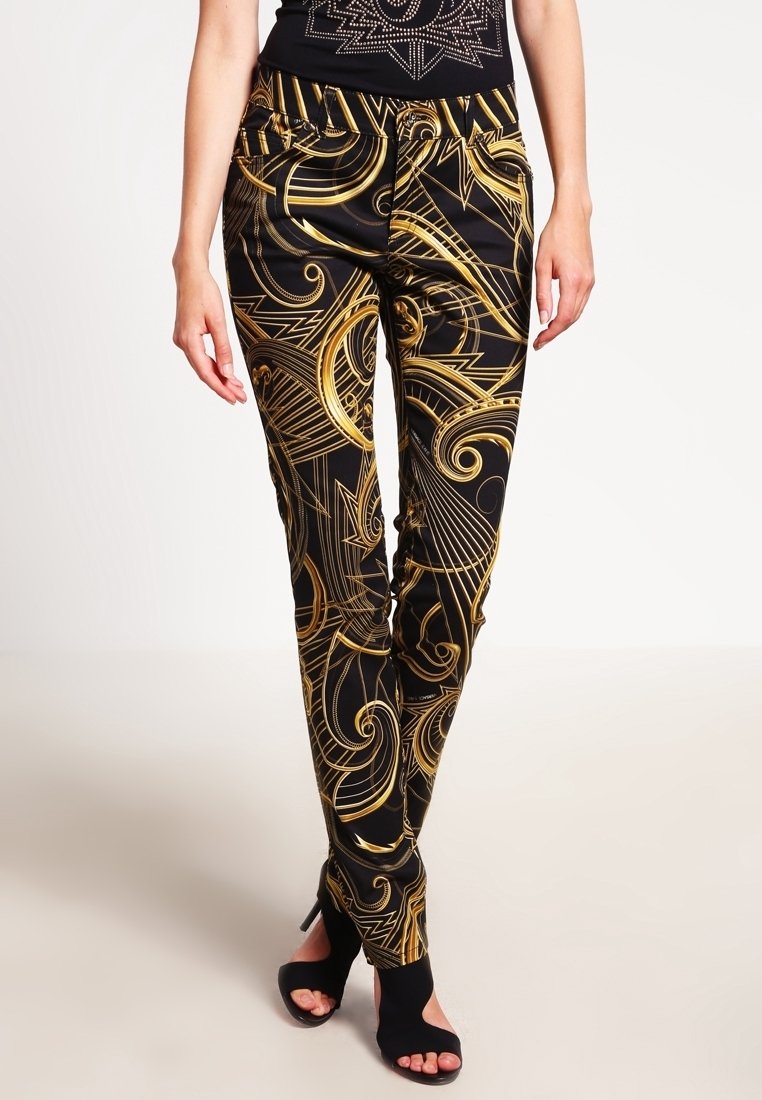 Pantalons noirs avec des motifs abstraits dorés en spirale, présentant un design ajusté et une taille élastique, associés à des talons ouverts noirs.