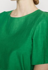 Nainen, jolla on päällään kirkkaan vihreä lyhythihainen paita, jossa on pyöreä kaula-aukko, näkyy niskaan asti ylävartalon alueelta tasaisella taustalla.