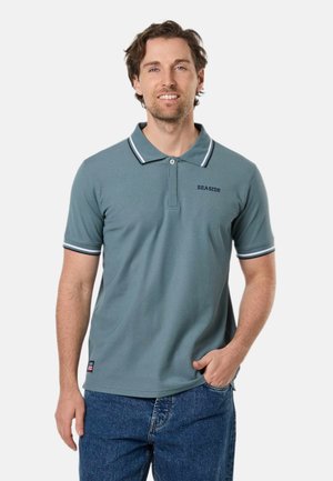 Uomo con capelli corti castani che indossa una polo turchese con il testo "SEASIDE" e jeans blu, in piedi con una mano in tasca.