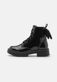 Bottine en cuir verni noir avec un bout rond, des lacets noirs, une semelle texturée et un détail en nœud à l'arrière. Talon plat et épais.