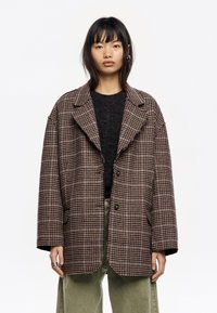 Blazer de cuadros marrón y beige de gran tamaño con solapa de muesca, dos botones y bordes desgastados, llevado sobre un suéter oscuro y pantalones verdes.