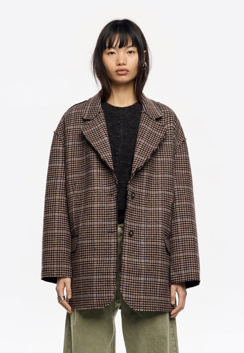 Blazer de cuadros marrón y beige de gran tamaño con solapa de muesca, dos botones y bordes desgastados, llevado sobre un suéter oscuro y pantalones verdes.
