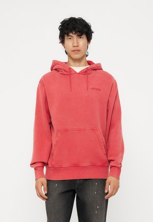 Joven con cabello rizado negro, vistiendo una sudadera roja con bolsillo delantero y vaqueros negros desgastados, de pie frente a un fondo blanco liso.
