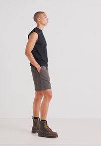 Musthave muströöm, hallid lühikesed püksid ja pruunid nööridega saapad kollaste aktsentidega, seistes profiilis tasase taustaga.