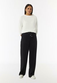 Witte textuur langemouwen top gecombineerd met high-waisted zwarte wijde broek en witte sneakers; heeft een strikdetail bij de taille.