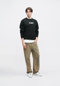 Μαύρο φούτερ με λαιμόκοψη και λευκό λογότυπο, σε συνδυασμό με λευκό πουκάμισο. Χαλαρές καφέ παντελόνες και λευκά αθλητικά παπούτσια ολοκληρώνουν την εμφάνιση.