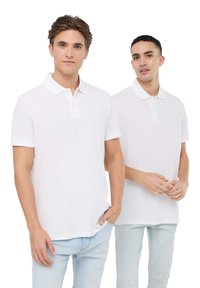 Due modelli indossano polo bianche semplici con maniche corte. Le polo hanno una chiusura con bottoni e sono abbinate a jeans di denim azzurro chiaro.