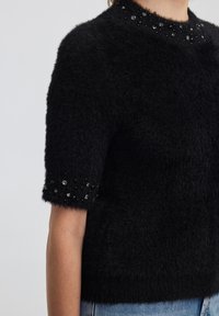 Czarna, puszysta bluzka z krótkim rękawem o teksturowanej, miękkiej powierzchni. Kołnierz i mankiety ozdobione czarnymi i przezroczystymi koralikami dla podkreślenia detali.