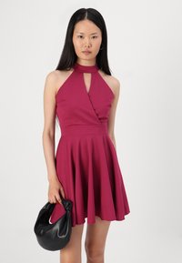 Robe halter ajustée magenta avec un détail découpé au niveau de la poitrine, tissu texturé et jupe évasée. Le modèle tient un sac en cuir noir.